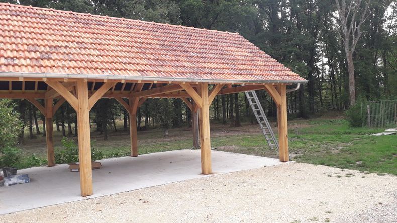 Pavillon en bois ouvert sur les côtés, avec un toit de tuiles rouges, situé sur une zone de gravier près des arbres.