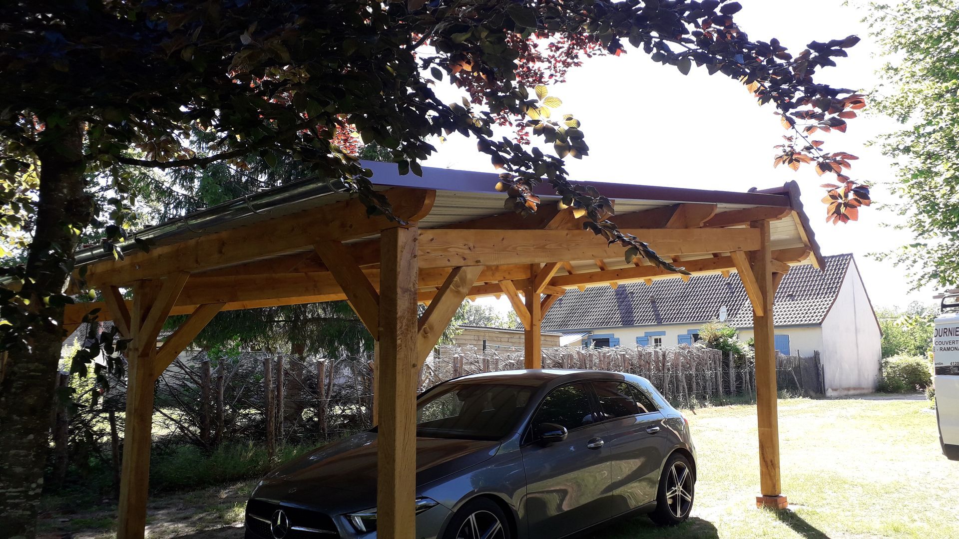 Un abri de voiture en bois abrite une voiture sombre dans une cour ensoleillée, avec des arbres.