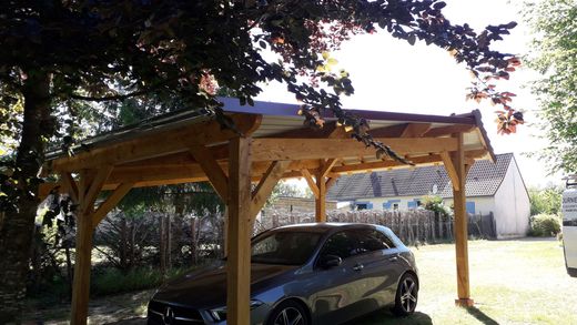 Un abri de voiture en bois abrite une voiture sombre dans une cour ensoleillée.