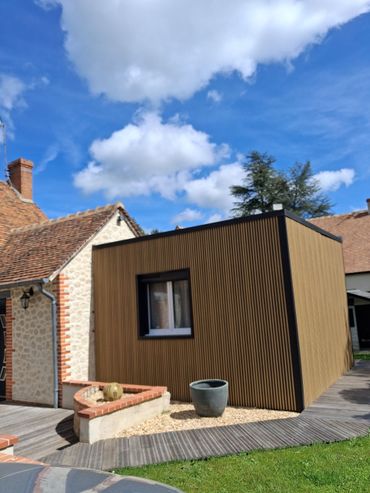 Petit bâtiment de jardin en bois avec une fenêtre, à côté d'une maison en briques et d'un patio sous un ciel bleu.