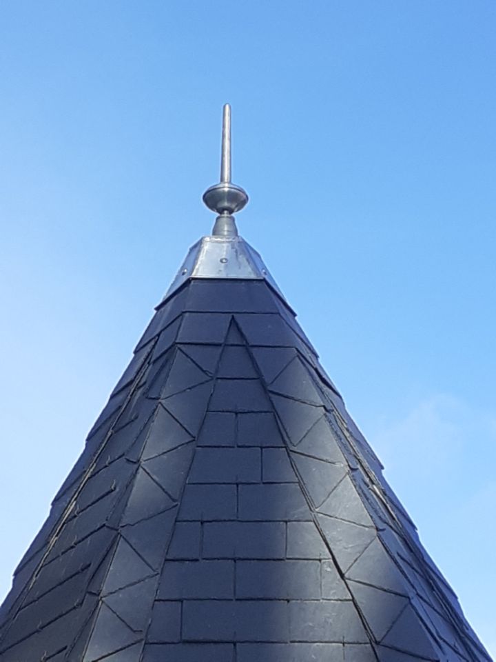 Toit pointu en tuiles sombres avec une flèche argentée se détachant sur un ciel bleu.