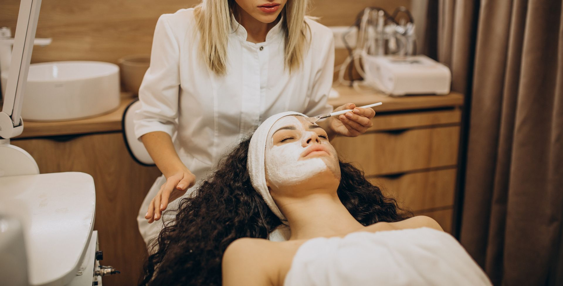 Una persona in uniforme bianca applica una maschera per il viso a un cliente sdraiato su una poltrona da trattamento all'interno di una spa.
