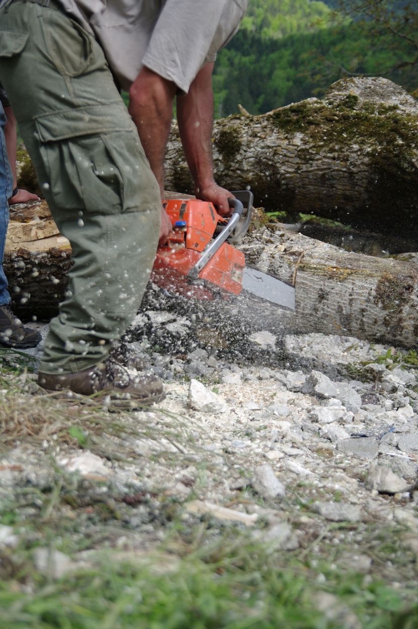 Une personne utilise une tronçonneuse pour couper une bûche sur le sol, de la sciure vole.