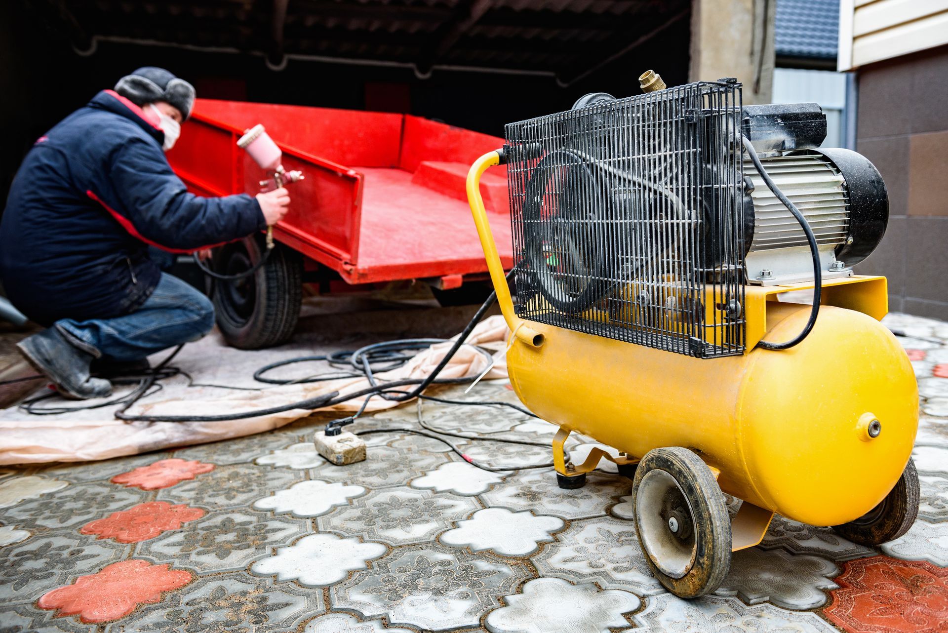Une personne utilise un pistolet à peinture pour peindre une remorque utilitaire rouge, alimentée par un compresseur d'air jaune, sur une terrasse en pierre.