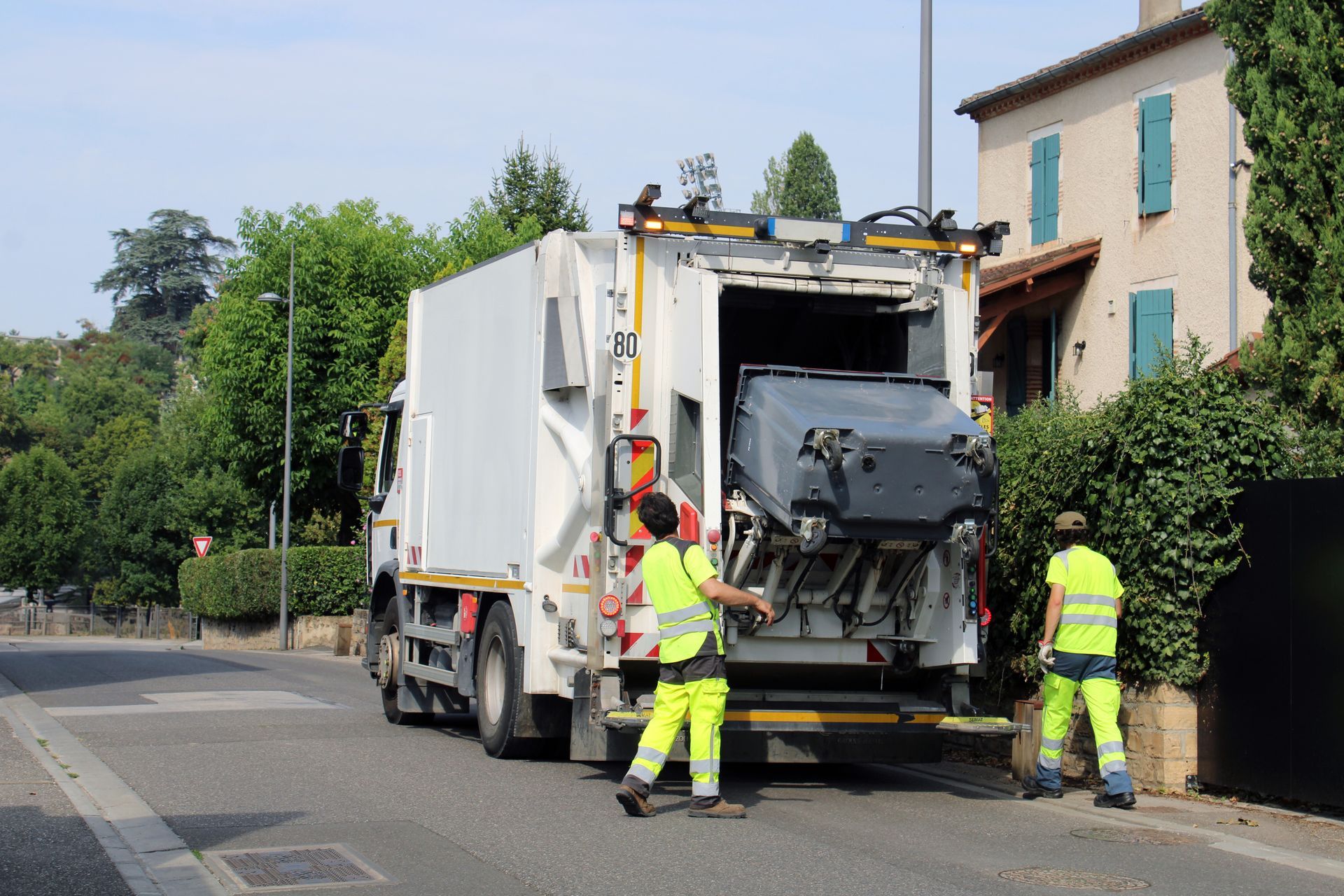Deux employés vêtus de gilets jaune vif haute visibilité manœuvrent un camion-poubelle pour vider un grand conteneur dans une rue ensoleillée de banlieue.