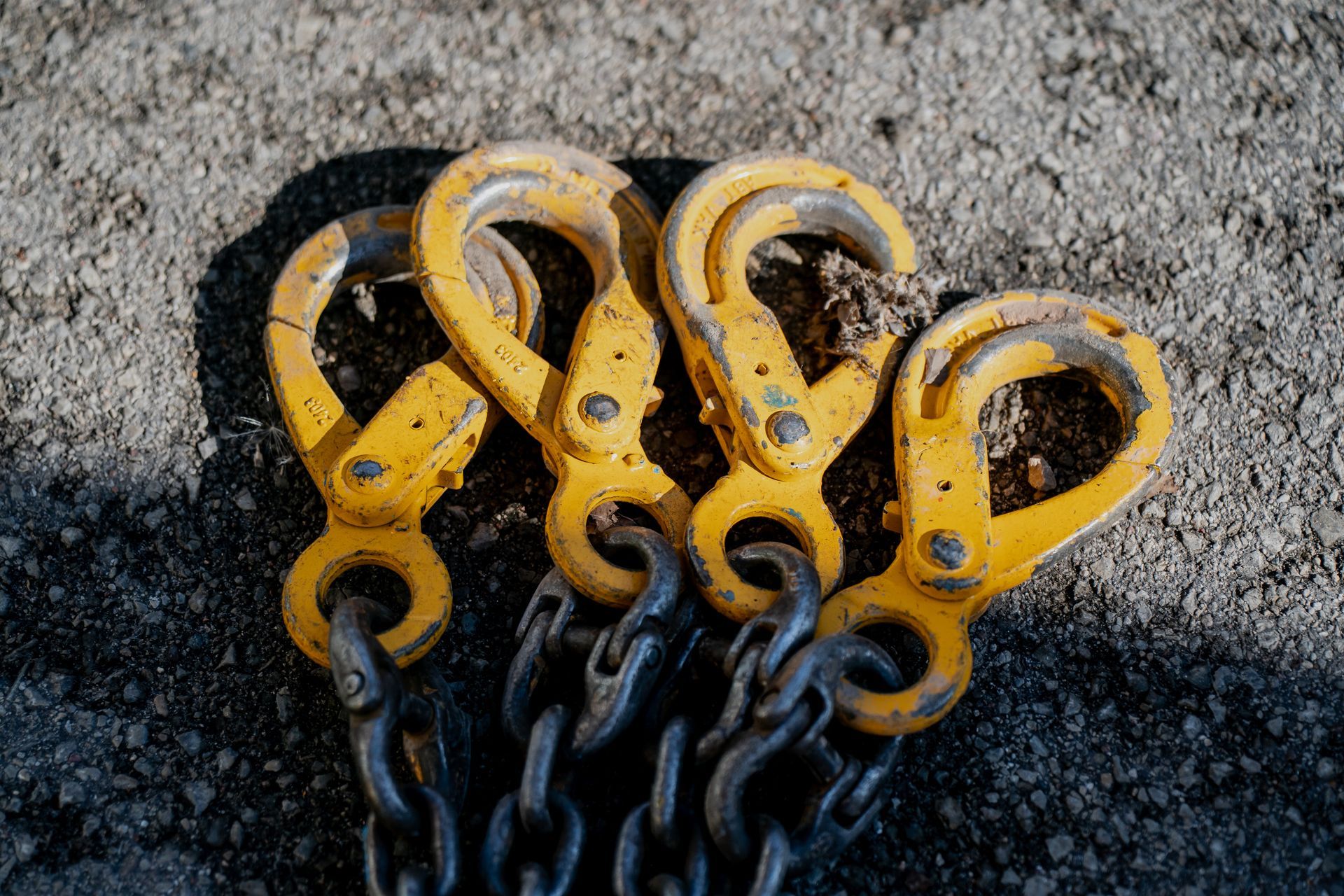 Quatre crochets métalliques jaunes, fixés à des chaînes sombres, sont disposés côte à côte sur une surface de gravier texturée.