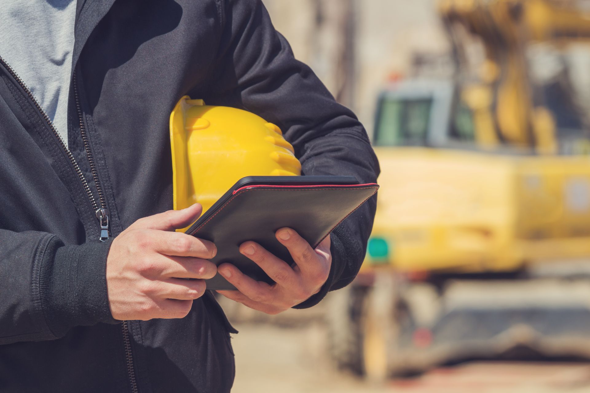 Une personne portant une veste sombre, tenant un casque de chantier jaune et une tablette, sur un chantier de construction avec des engins lourds.