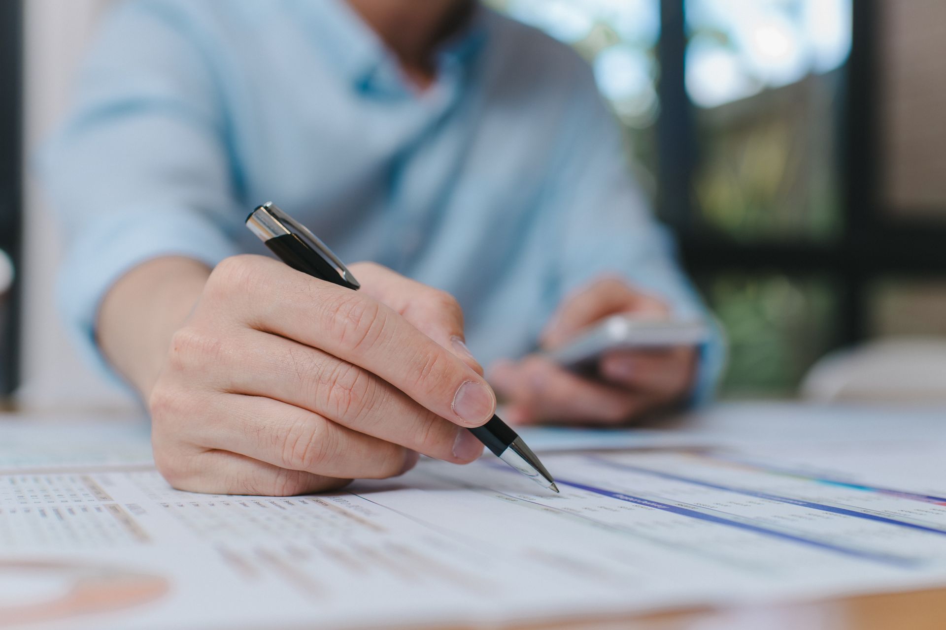 Une personne vêtue d'une chemise bleu clair écrit sur un document financier avec un stylo tout en tenant un smartphone.