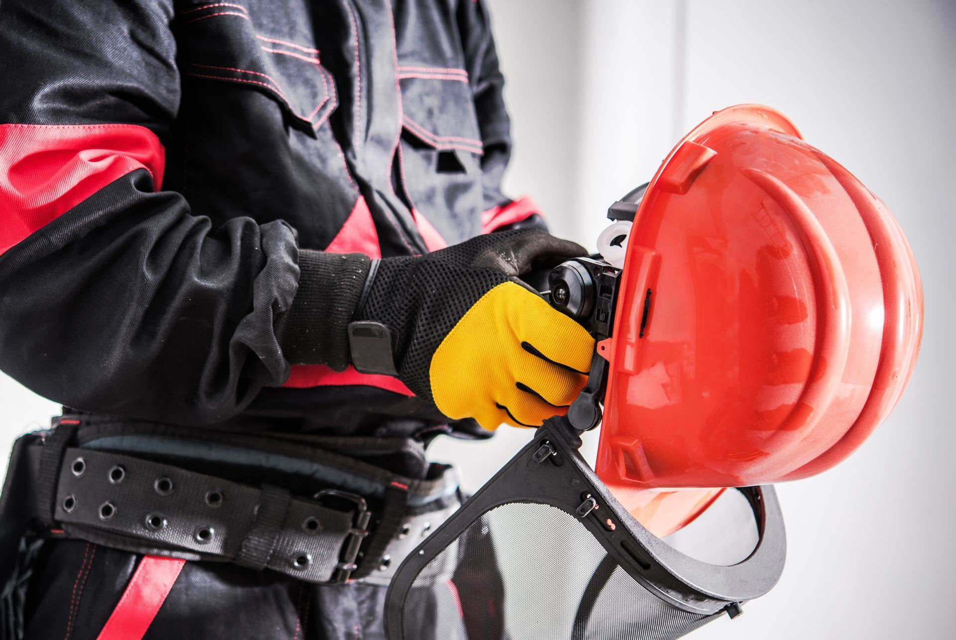 Un ouvrier du bâtiment en uniforme noir et rouge et gants jaunes tient un casque de chantier muni d'une visière de protection en maille.