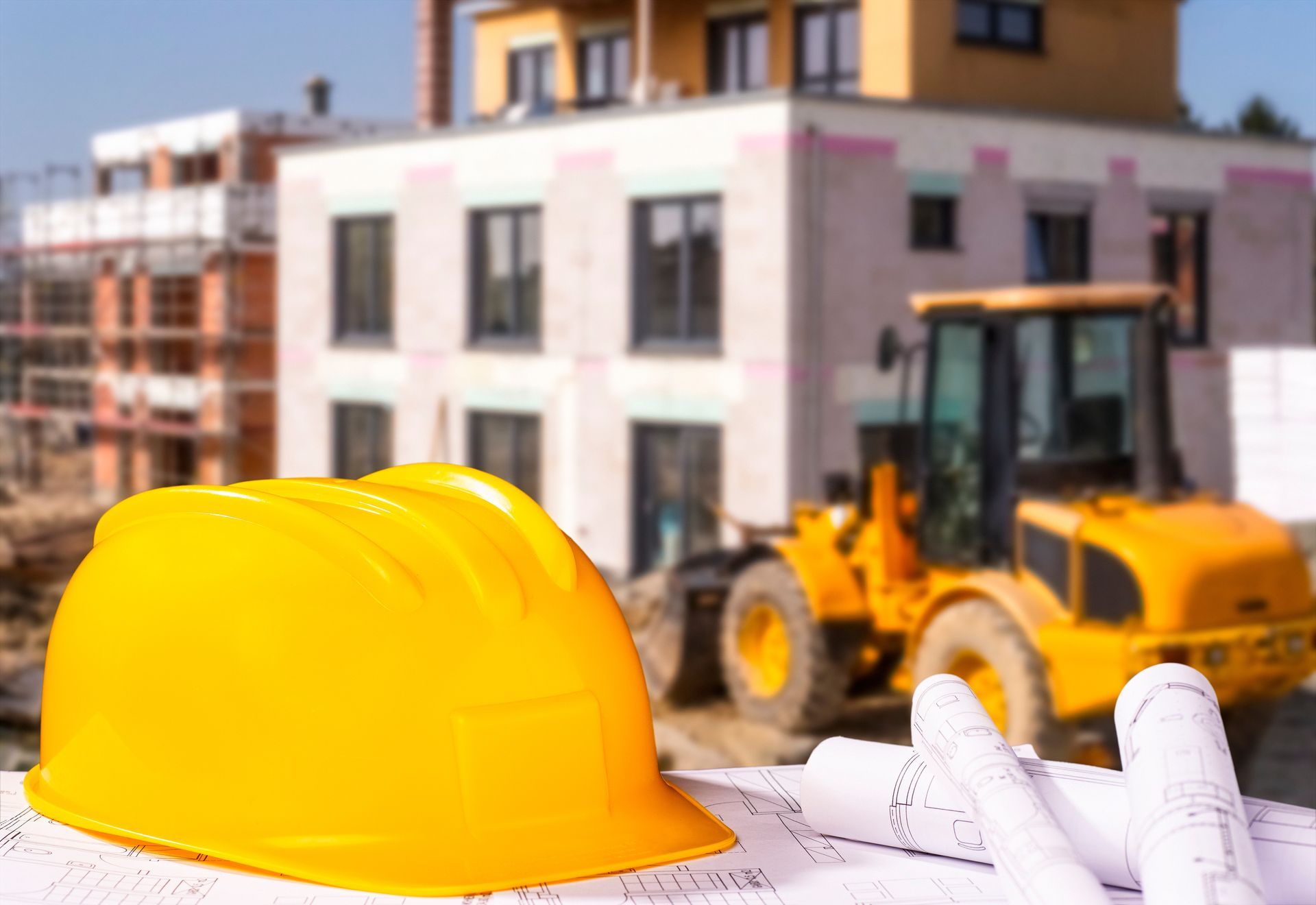 Un casque de chantier jaune est posé sur des plans de construction, avec une chargeuse jaune et une maison en construction à l'arrière-plan.
