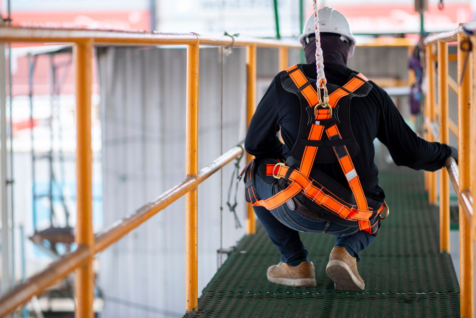 Un ouvrier du bâtiment, portant un casque blanc et un harnais de sécurité orange, est accroupi sur une plateforme métallique aux rambardes jaunes.