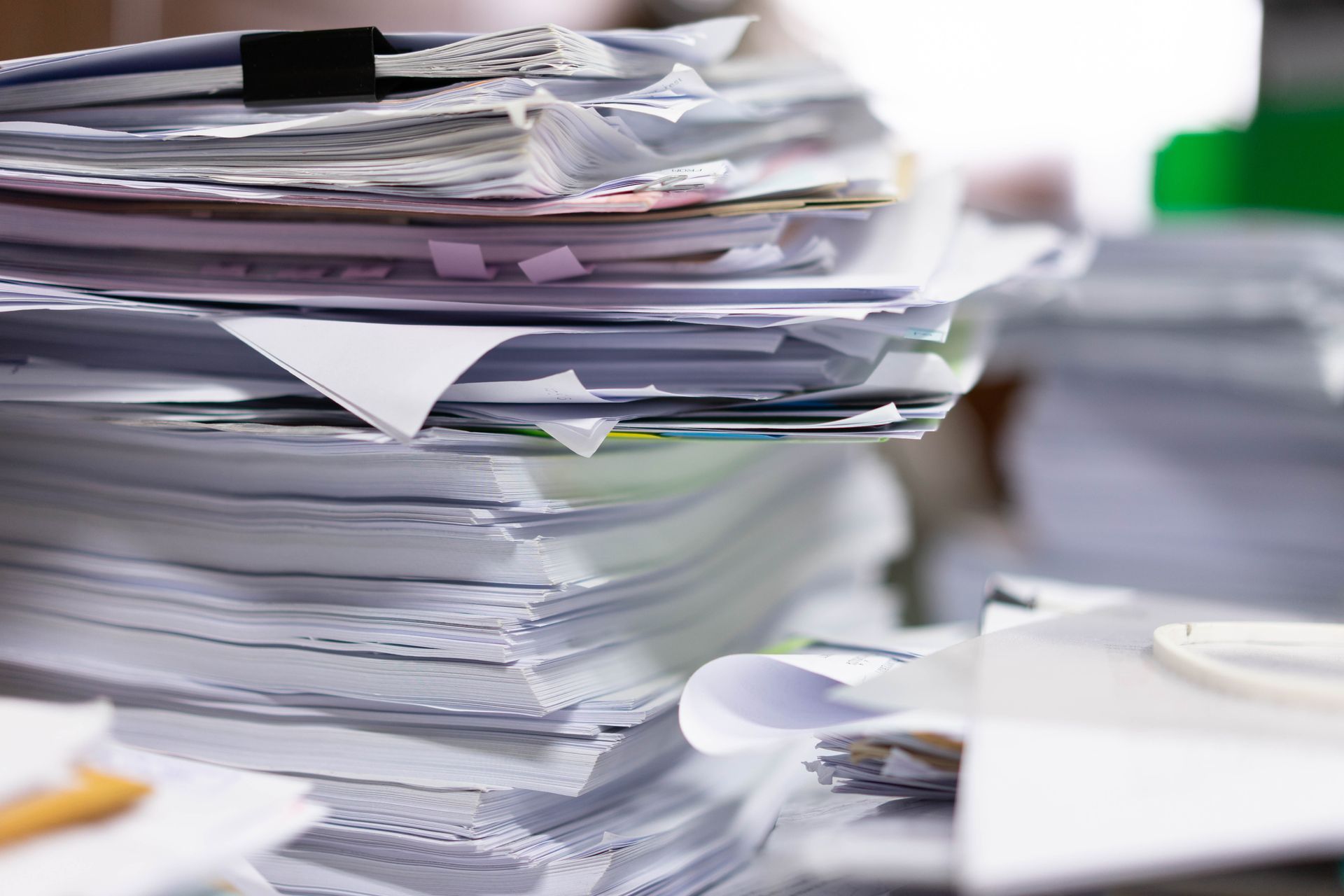 Une grosse pile de documents blancs en désordre sur un bureau, avec une pince à reliure noire au sommet.
