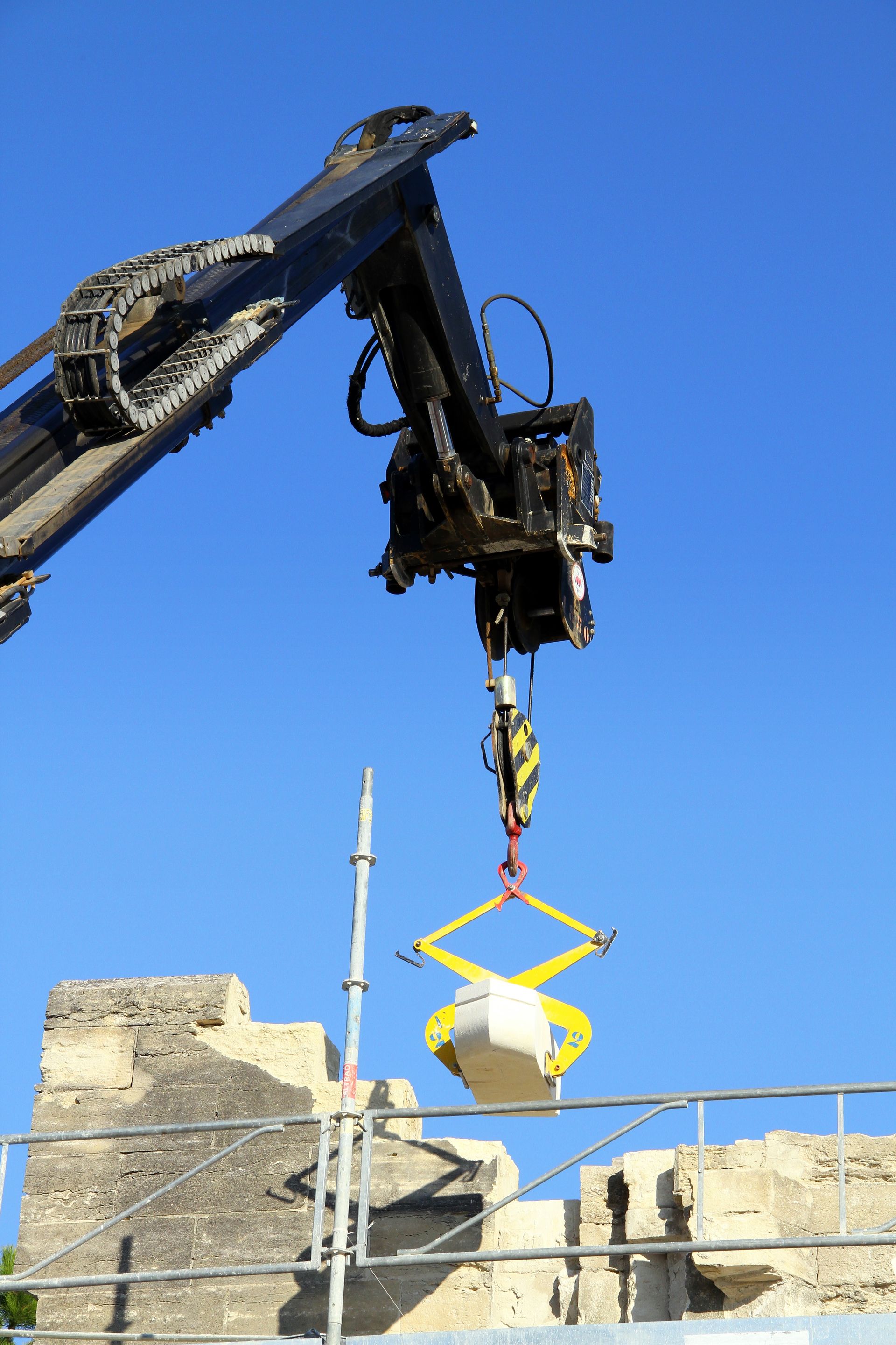 Sur fond de ciel bleu, le bras d'une grue mécanique soulève un lourd bloc de pierre à l'aide d'une pince de levage jaune en forme de ciseaux.