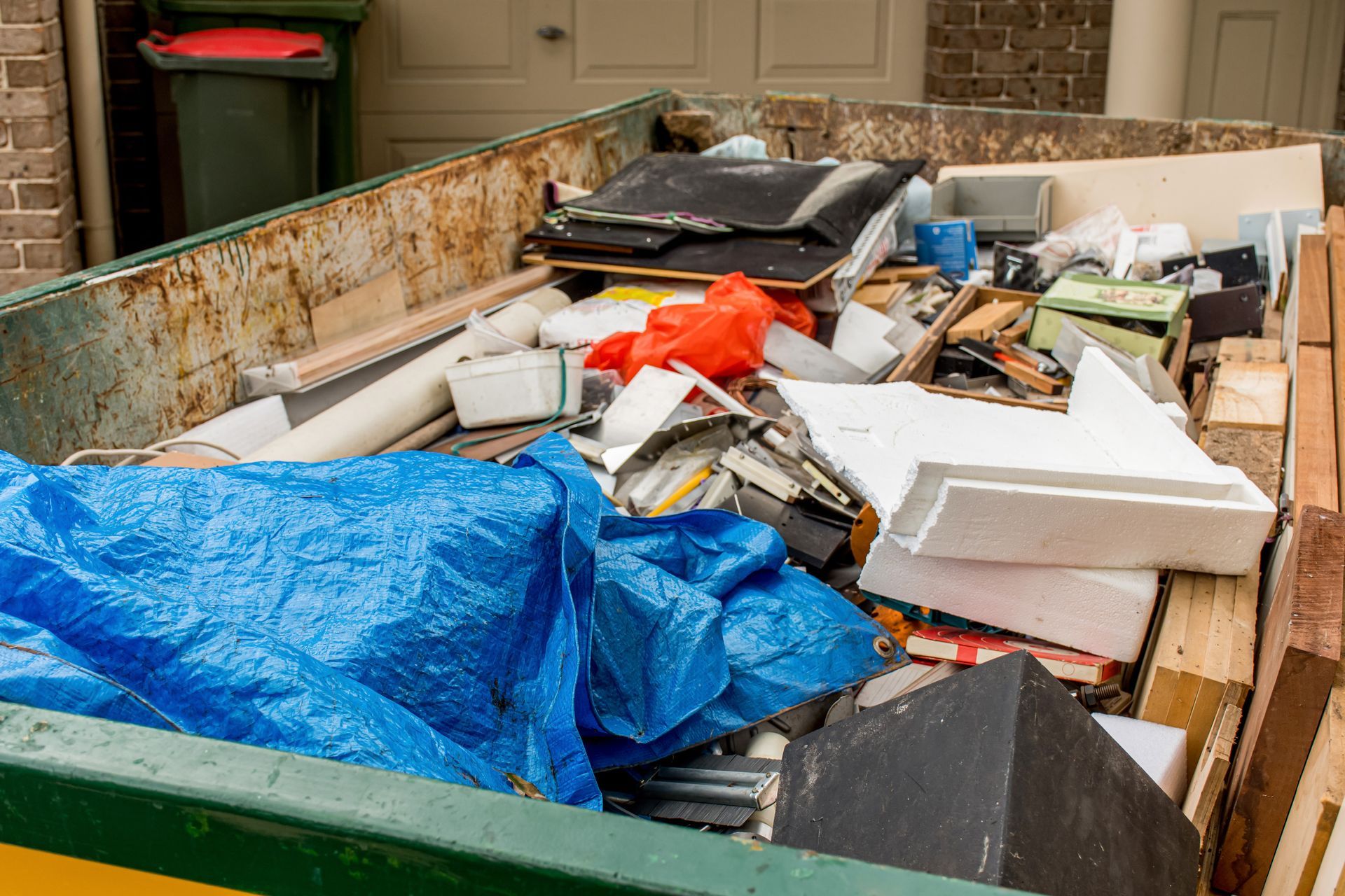 Une benne remplie de déchets ménagers divers, de débris de construction, d'une bâche bleue et de blocs de mousse blanche.