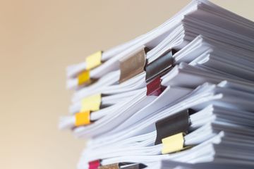Une pile de documents en papier blanc, maintenus par des pinces à reliure métalliques de différentes couleurs, se détache sur un fond clair uni.