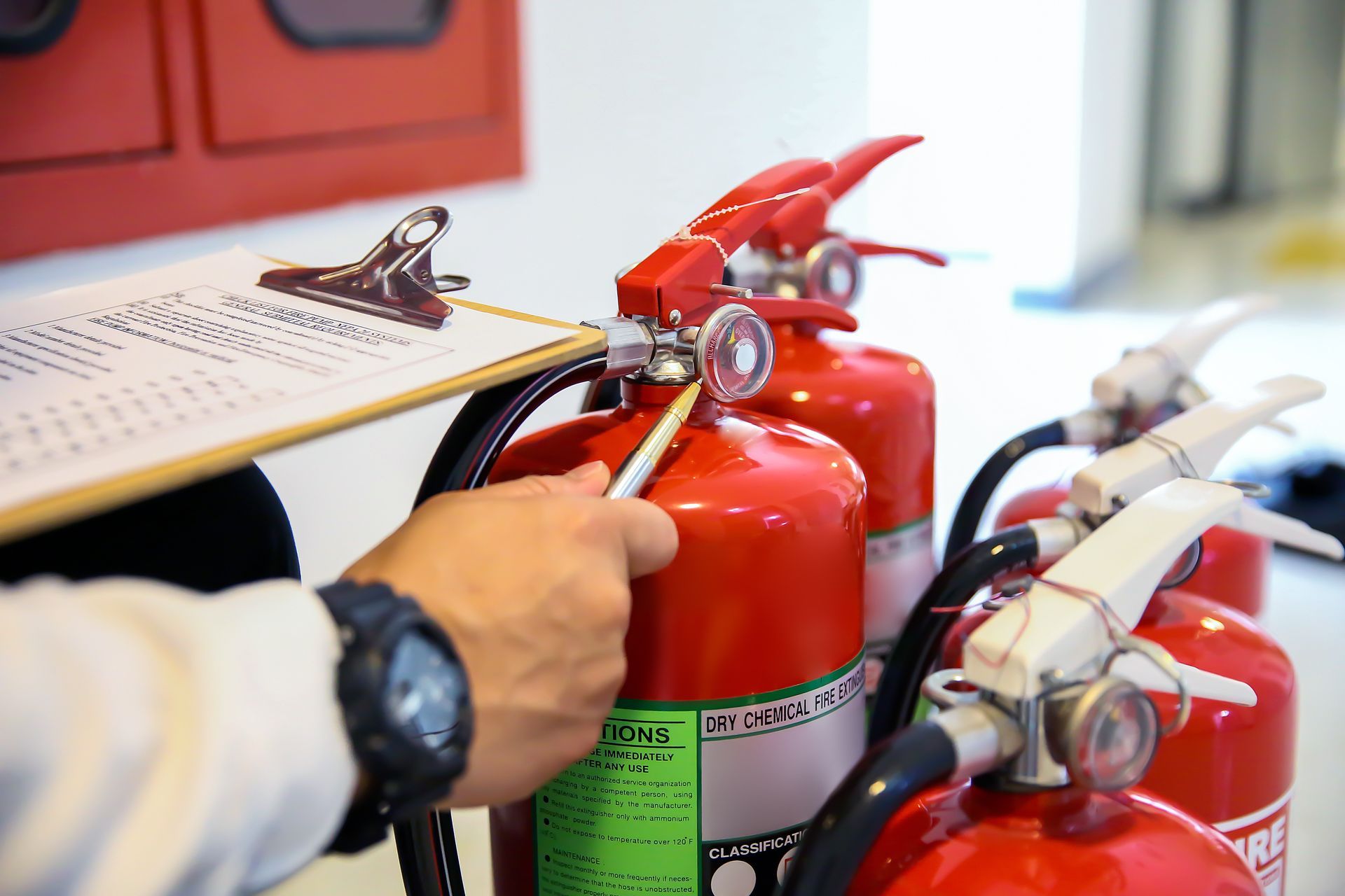 Une main tient un stylo pour inspecter un extincteur rouge tout en consultant une liste sur un bloc-notes lors d'une opération de maintenance de sécurité.