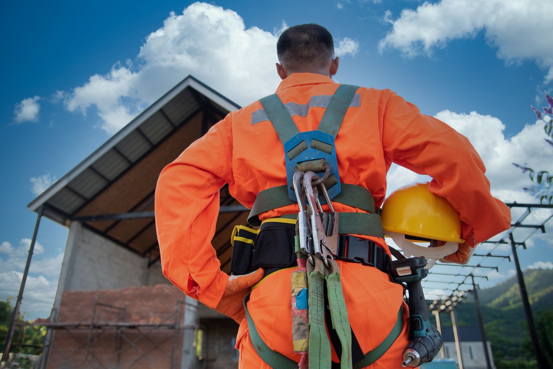 Un ouvrier du bâtiment en combinaison orange et harnais de sécurité, tenant un casque jaune, se tient devant un chantier.