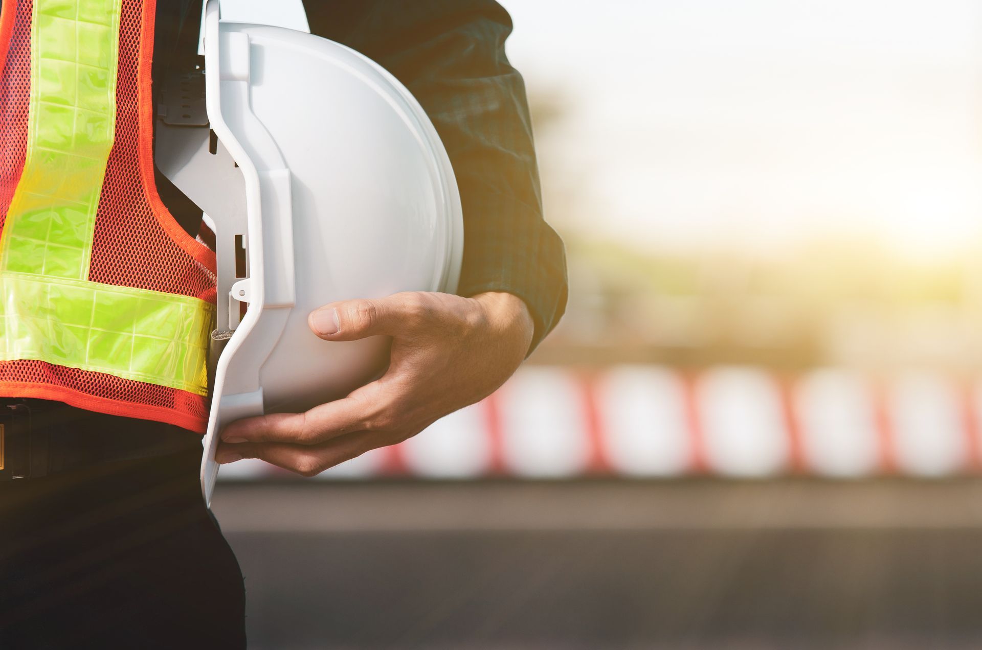 Une personne portant un gilet haute visibilité tient un casque de chantier blanc près d'un chantier de construction flou.
