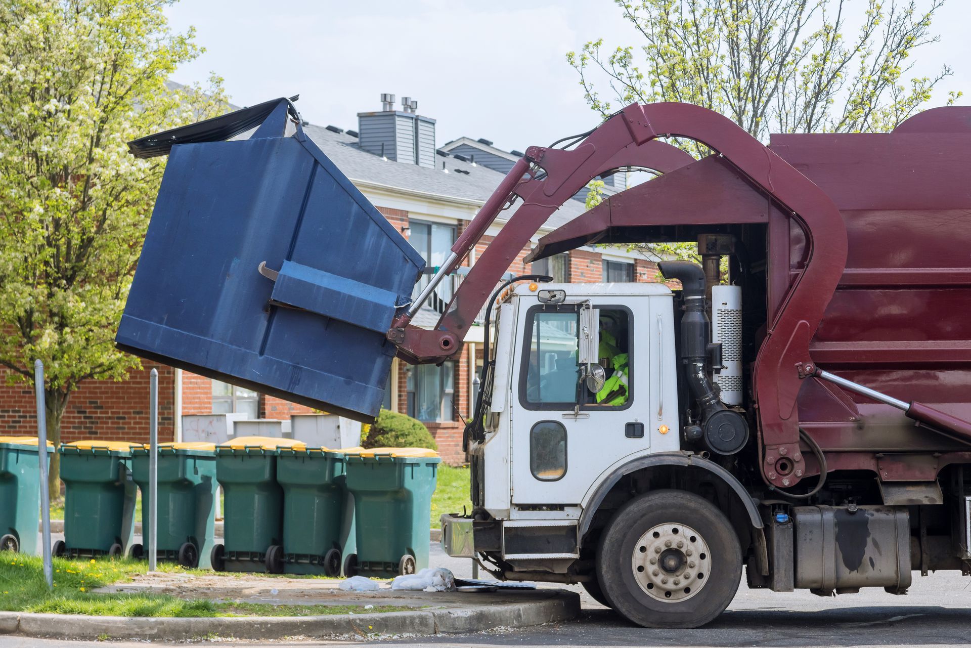 Un camion-poubelle bordeaux utilise un bras mécanique pour soulever un grand conteneur bleu dans une rue résidentielle ensoleillée de banlieue.