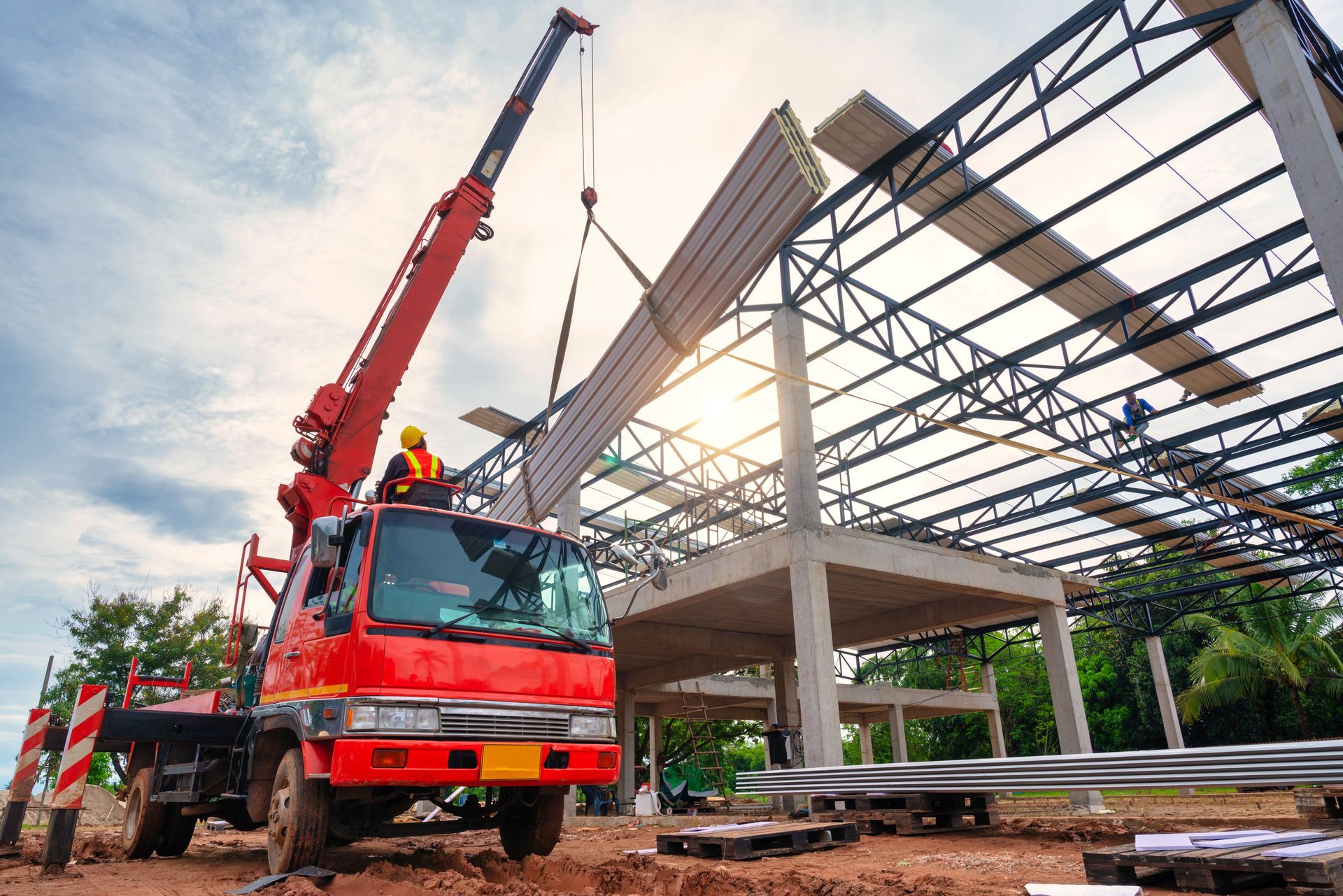 Une grue rouge soulève une poutre en béton sur un chantier de construction où la structure métallique d'un bâtiment est partiellement édifiée.
