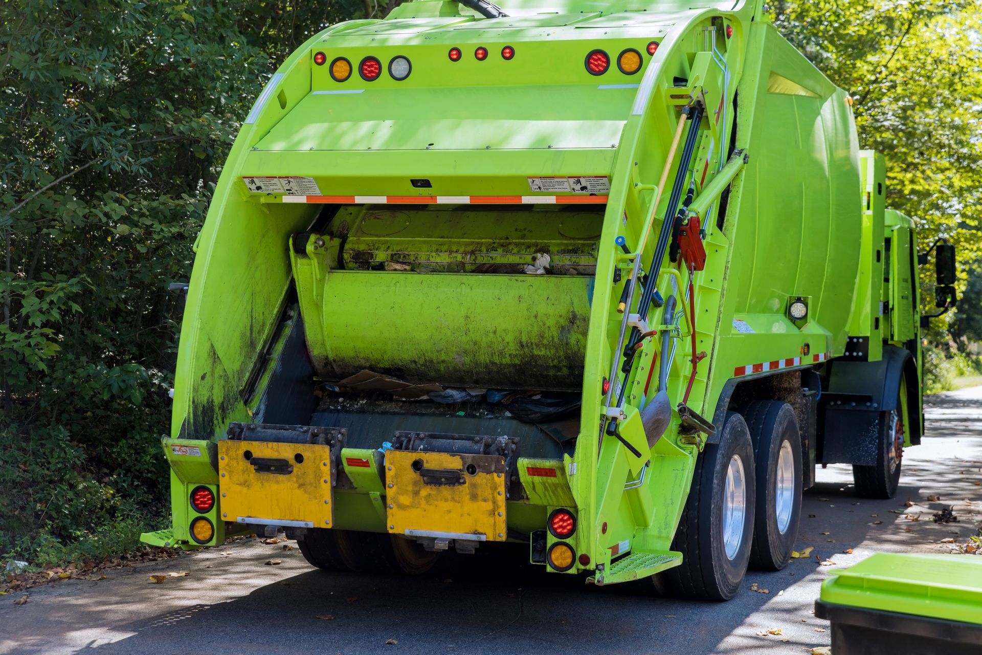 Un camion-poubelle vert citron vif est stationné sur une route bordée d'arbres, son mécanisme de chargement arrière étant visible.