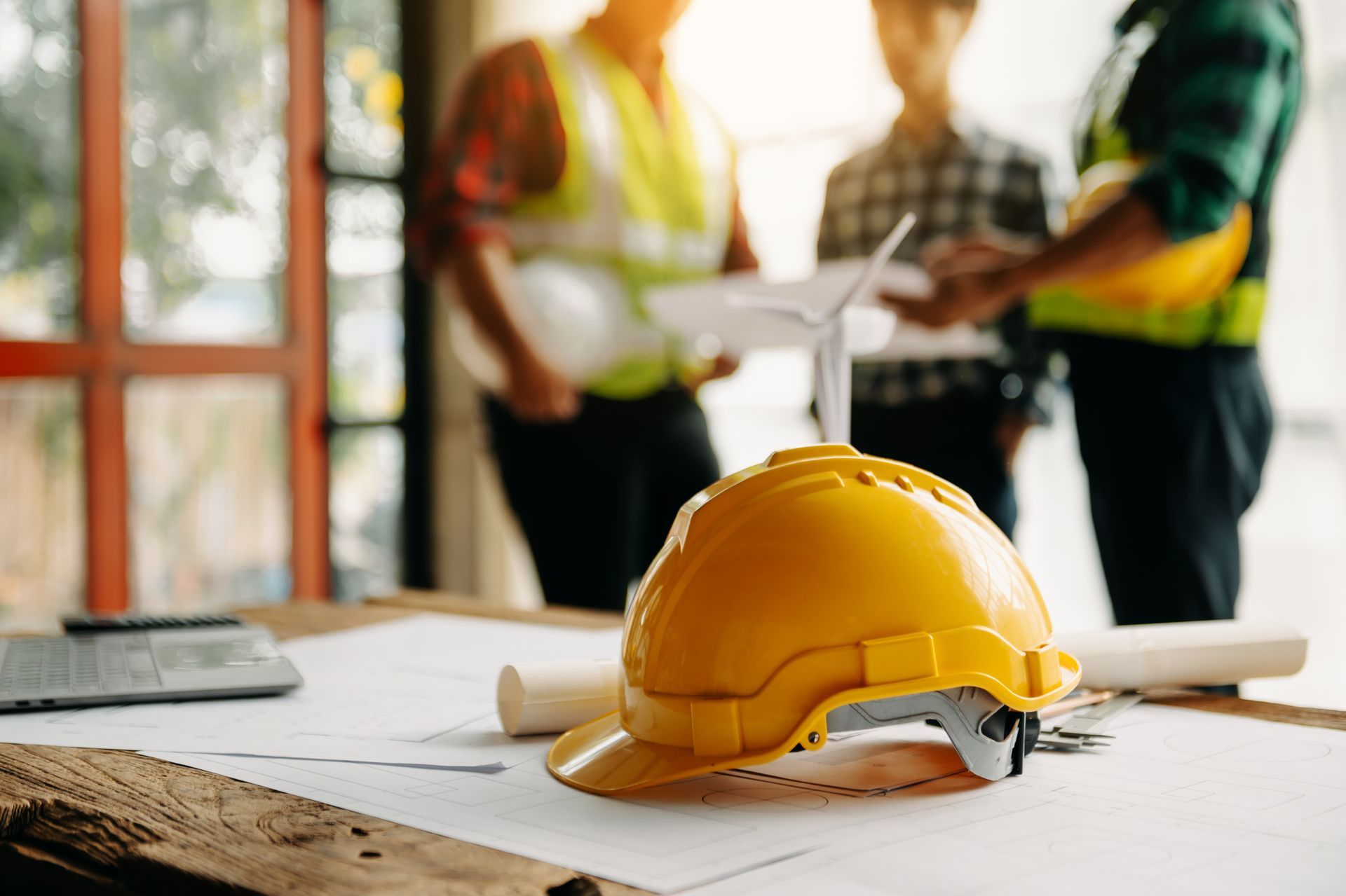 Au premier plan, un casque de chantier jaune repose sur des plans posés sur un bureau, tandis qu'à l'arrière-plan, trois professionnels du bâtiment se devinent de façon floue.