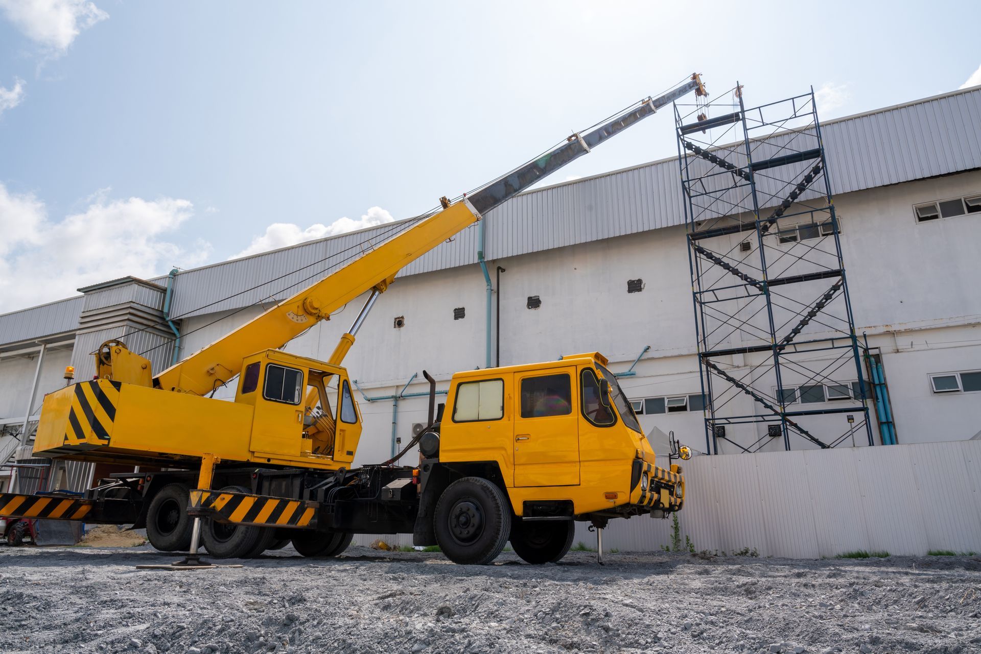 Une grue jaune montée sur camion est stationnée sur du gravier, sa flèche déployée vers une structure d'échafaudage métallique près d'un bâtiment.