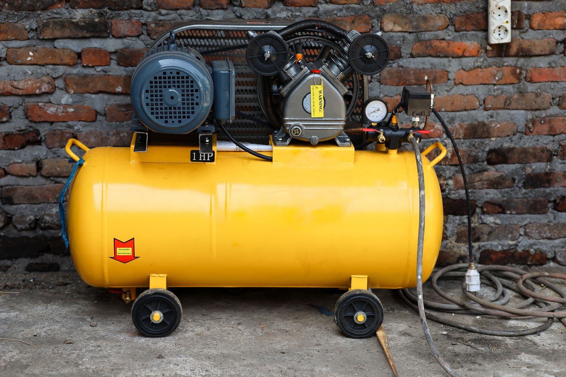Un compresseur d'air portable jaune vif, doté d'un moteur électrique et d'un réservoir gris, est posé sur un sol en briques, contre un mur en briques.