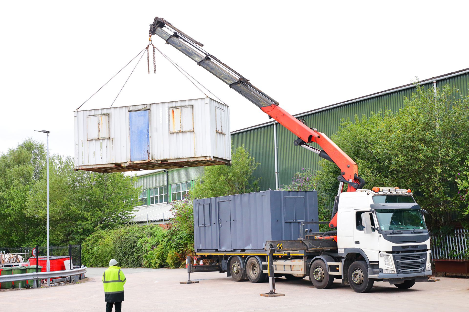 Un camion-grue soulève un module de bureau blanc au-dessus d'un conteneur gris foncé dans une cour extérieure, un ouvrier se trouvant à proximité.