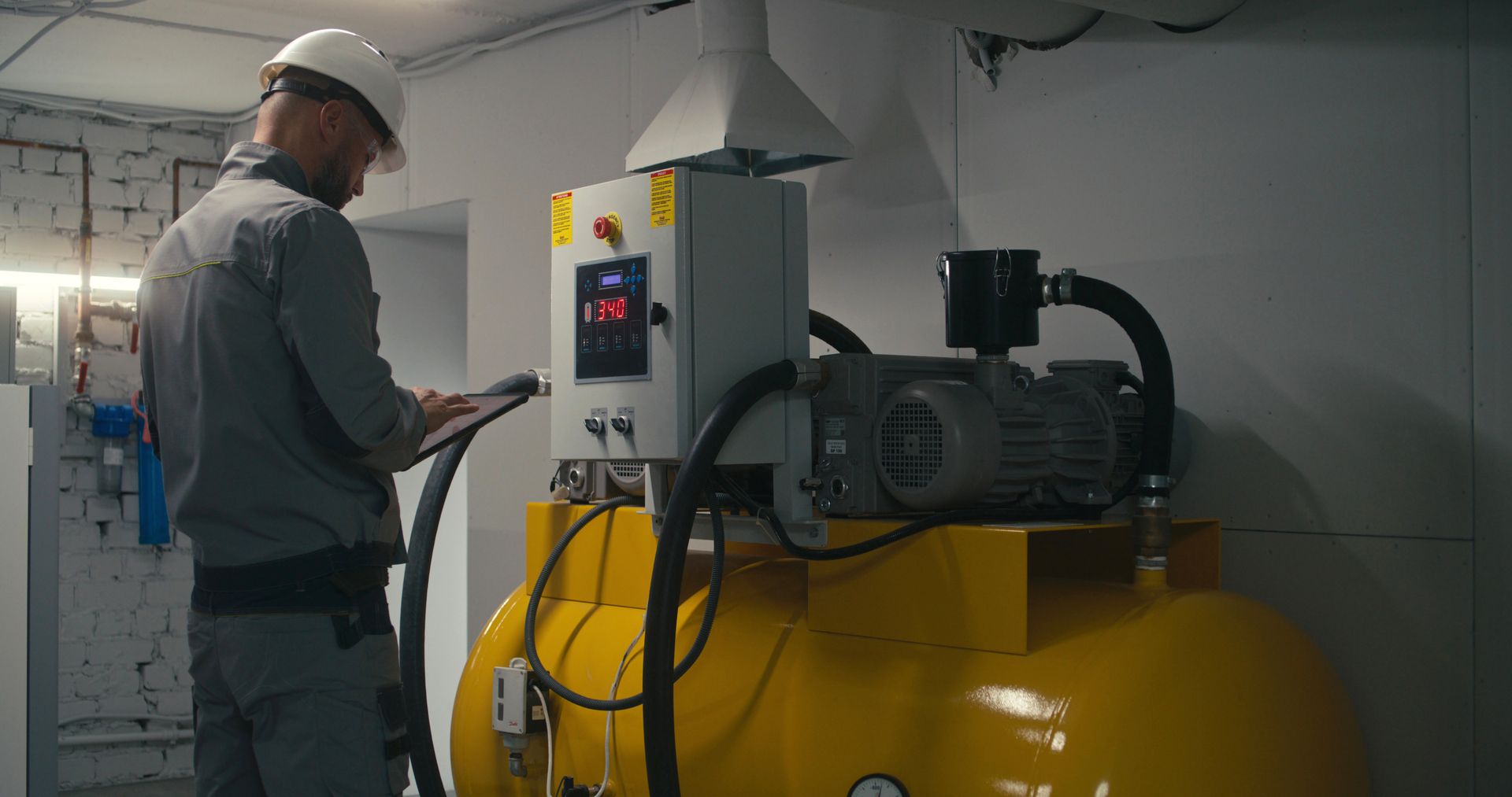 Dans un local technique, une personne en tenue de travail et casque de chantier consulte des données sur une tablette à côté d'un compresseur industriel jaune.