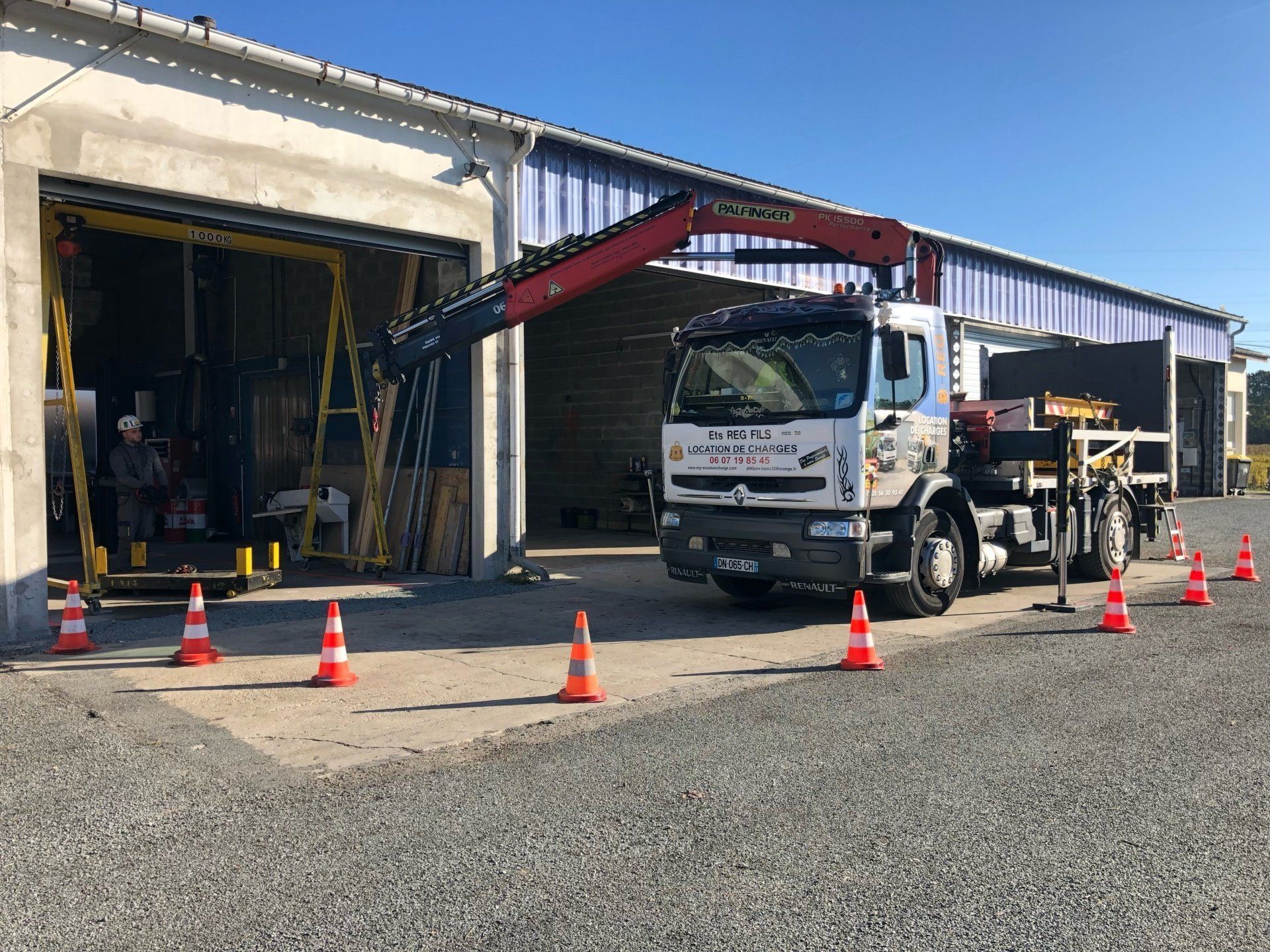 Un camion blanc équipé d'une grue rouge est stationné devant un entrepôt, entouré de cônes de signalisation orange sur un terrain gravillonné.