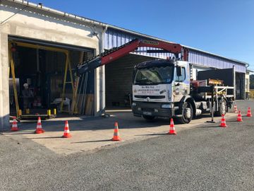 Un camion blanc avec une grue rouge est garé devant un entrepôt, encadré par des cônes de signalisation orange sur un terrain gravillonné.