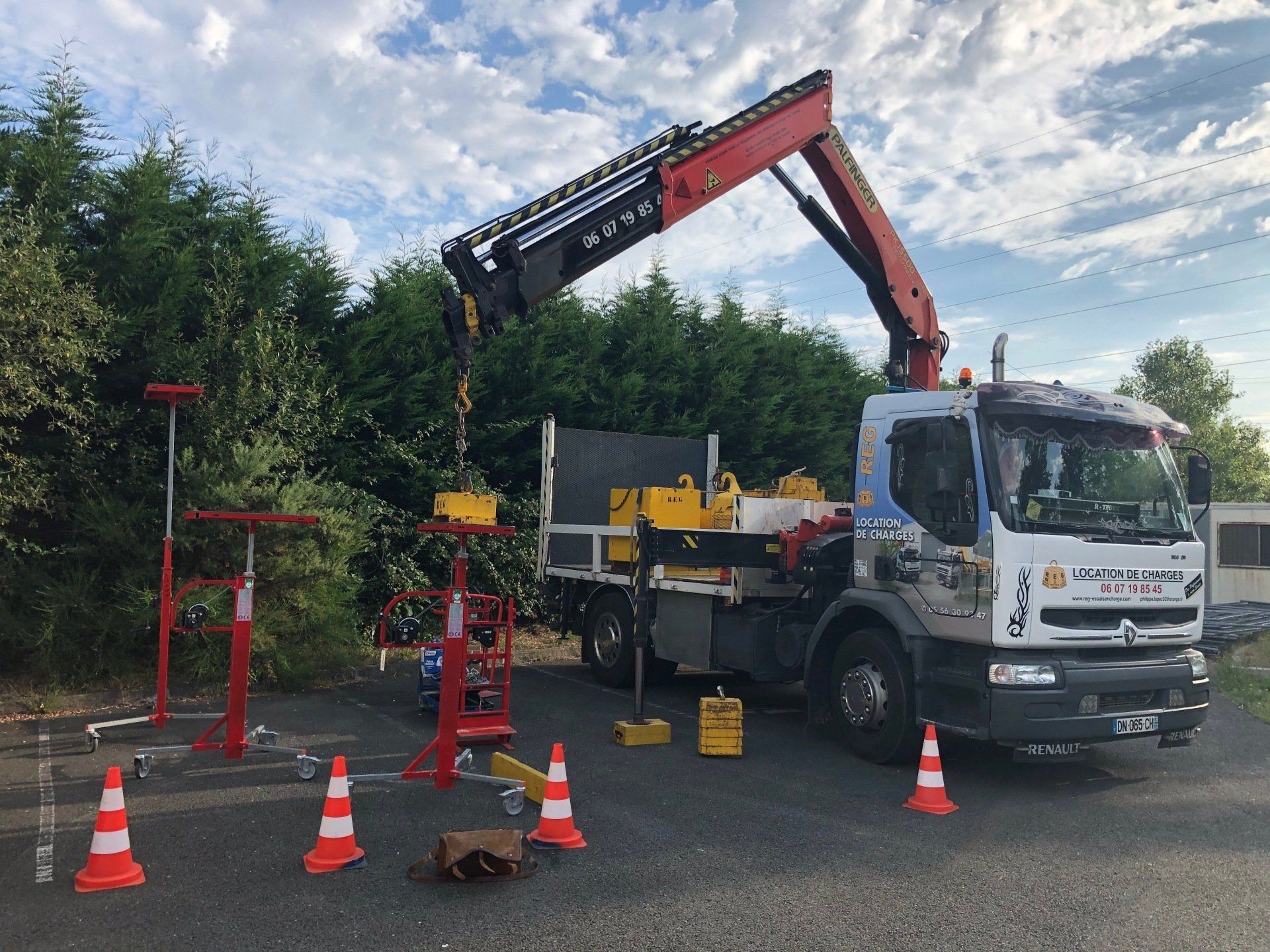 Un camion utilitaire blanc équipé d'une grue rouge est stationné à l'extérieur, entouré de cônes de signalisation orange et d'engins de levage mobiles.