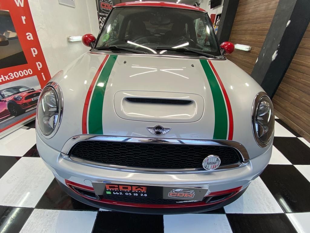Un Mini Cooper blanco con rayas rojas, verdes y blancas en el capó está estacionado en un garaje.