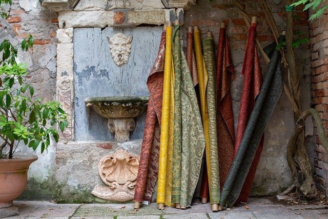 Bunte Stoffrollen lehnen an einer antiken Steinmauer mit einem Brunnen in einem Außenhof.