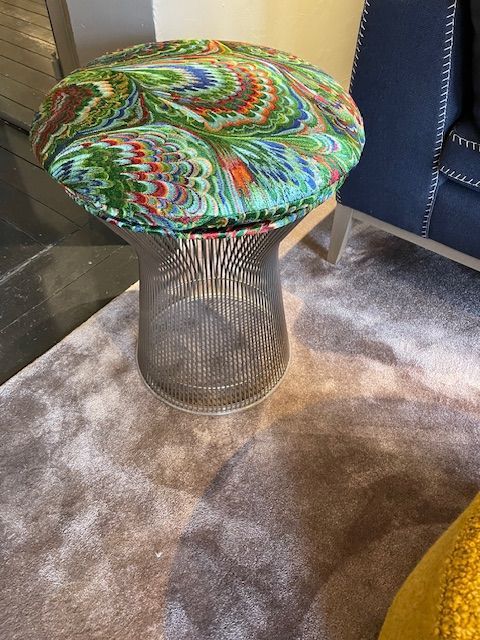 Ein silberner Drahthocker mit einem runden, farbenfrohen Sitz aus marmoriertem Stoff auf einem braunen Teppich.