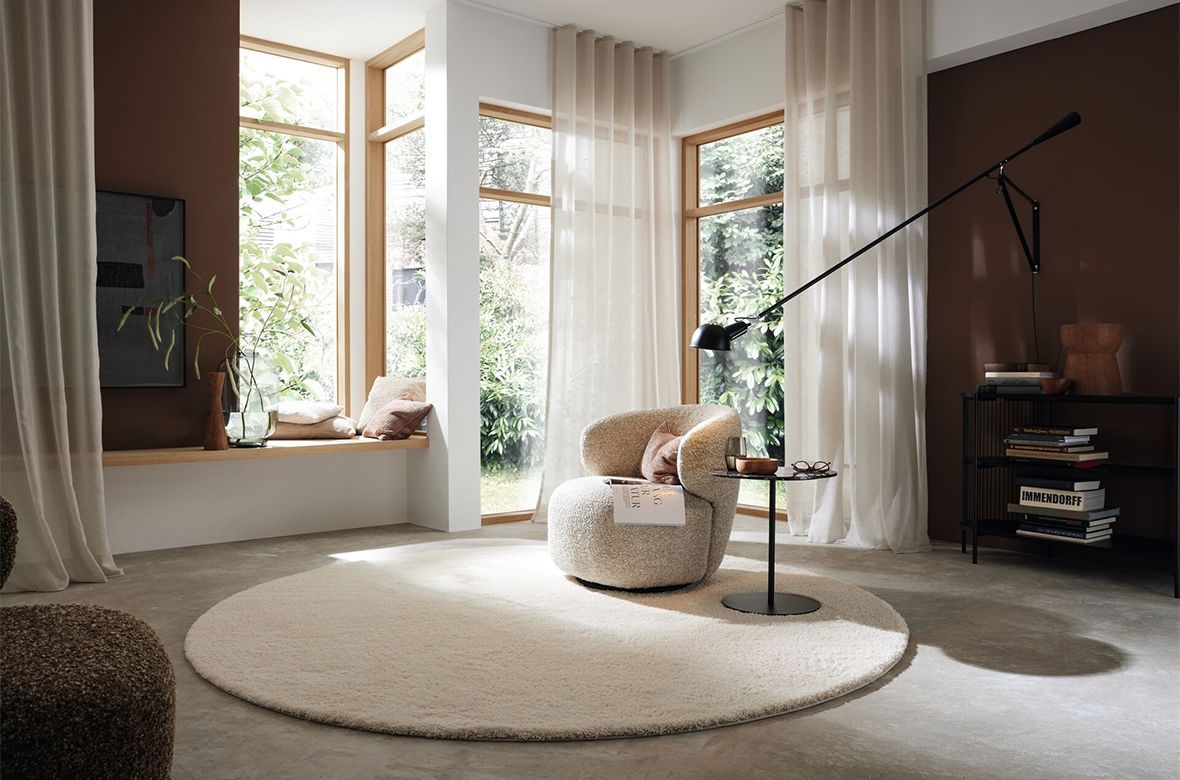 Gemütliches Wohnzimmer mit cremefarbenem Teppich, Sessel, transparenten Vorhängen und einer Leseecke.