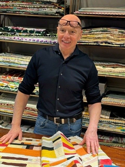 Eine lächelnde Person in dunklem Hemd und Jeans steht hinter einem Tisch