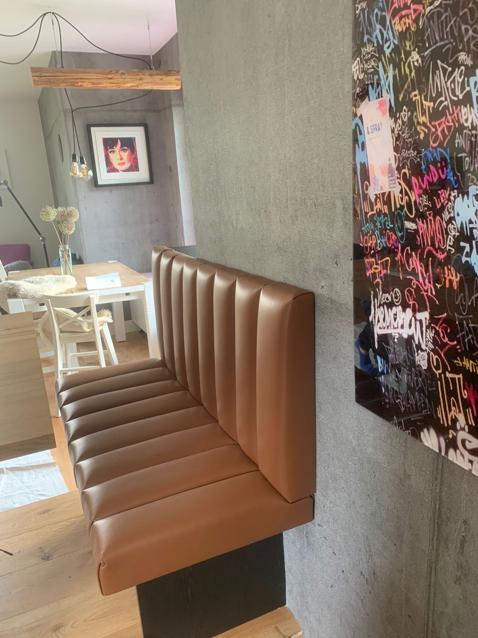 Eine braune Lederbank ist an einer grauen Wand befestigt, daneben befinden sich farbenfrohe Kunstwerke und ein Essbereich.