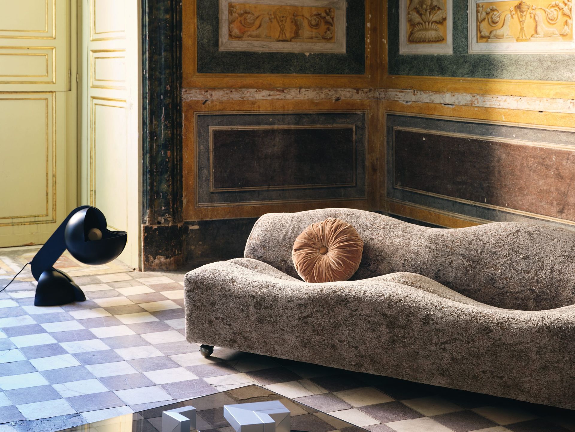 Schwarze Lampe neben einem strukturierten grauen Sofa mit einem runden Kissen. Schachbrettmusterboden, reich verzierte Wände.