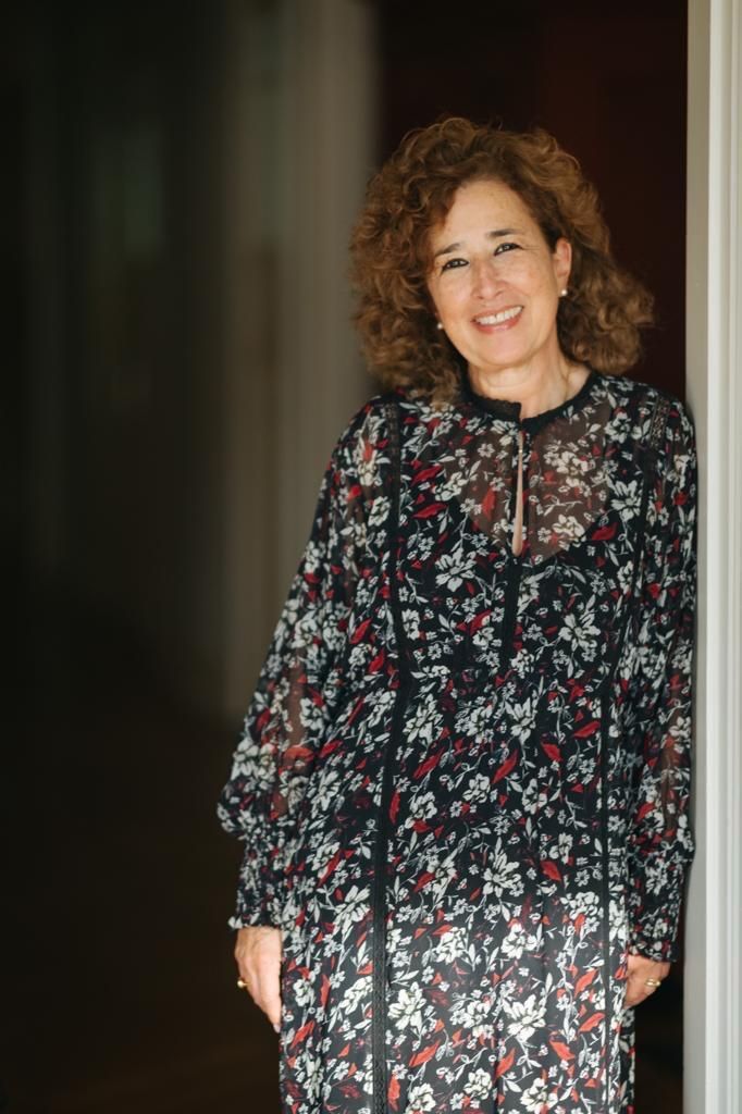 Eine Frau mit lockigem braunem Haar, die ein Kleid mit Blumenmuster trägt, lächelt, während sie sich an einen Türrahmen lehnt.