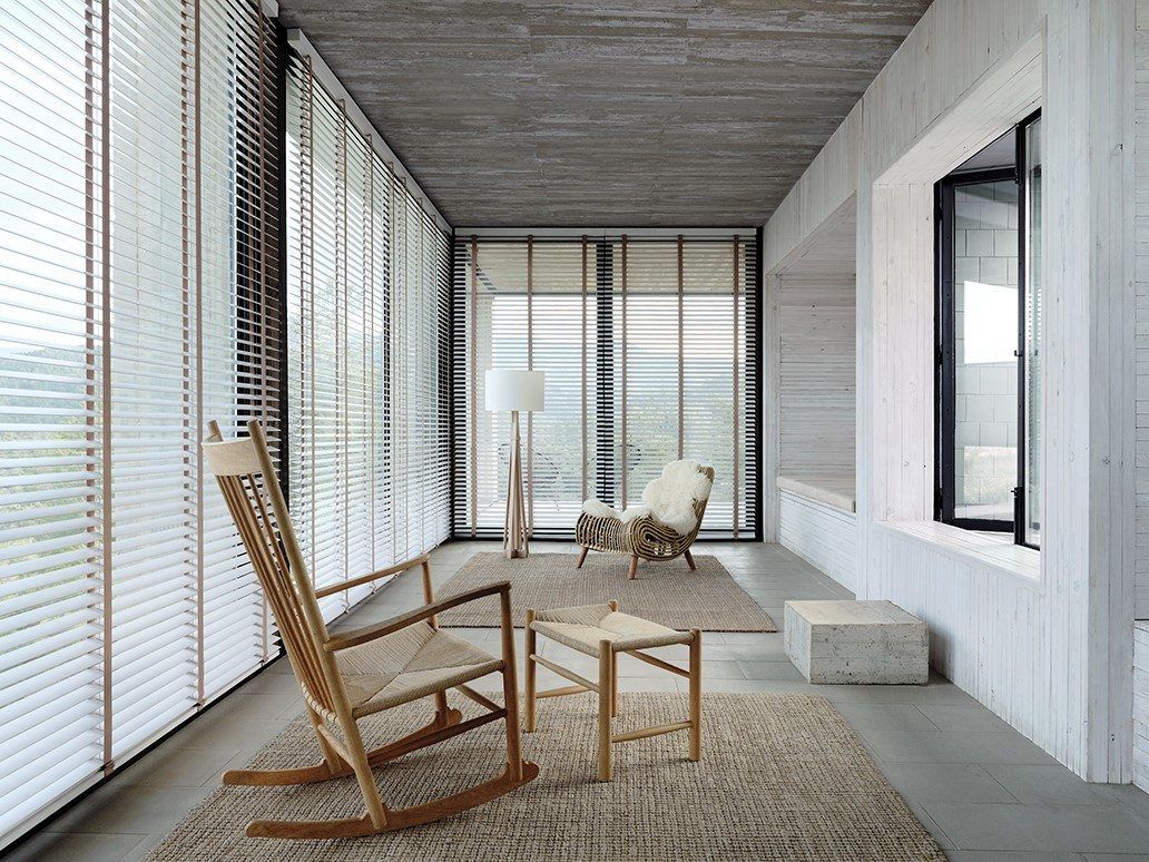 Innenansicht eines minimalistisch eingerichteten Zimmers mit Schaukelstuhl, Teppich und strukturierten Fensterdekorationen.