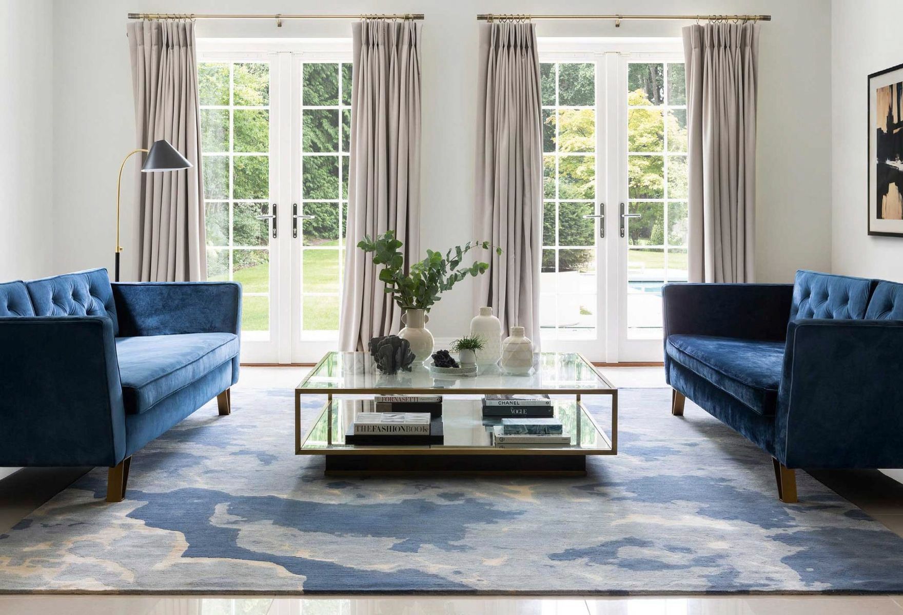 In einem hellen Raum stehen sich zwei blaue Samtsofas an einem gläsernen Couchtisch auf einem blau-weißen Teppich gegenüber