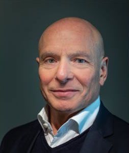 Glatzköpfiger Mann, lächelnd, der eine dunkle Jacke über einem weißen Hemd mit Kragen trägt.