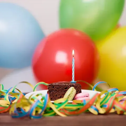 Un pastel de cumpleaños con una vela encendida en un plato con globos en el fondo.