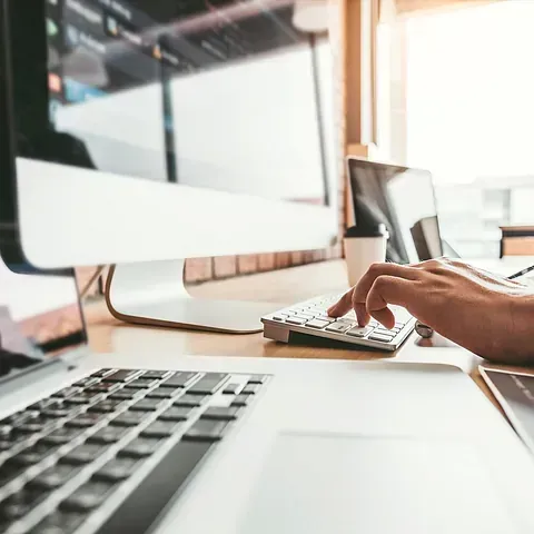 Una persona está escribiendo en un teclado portátil frente a un monitor de computadora.