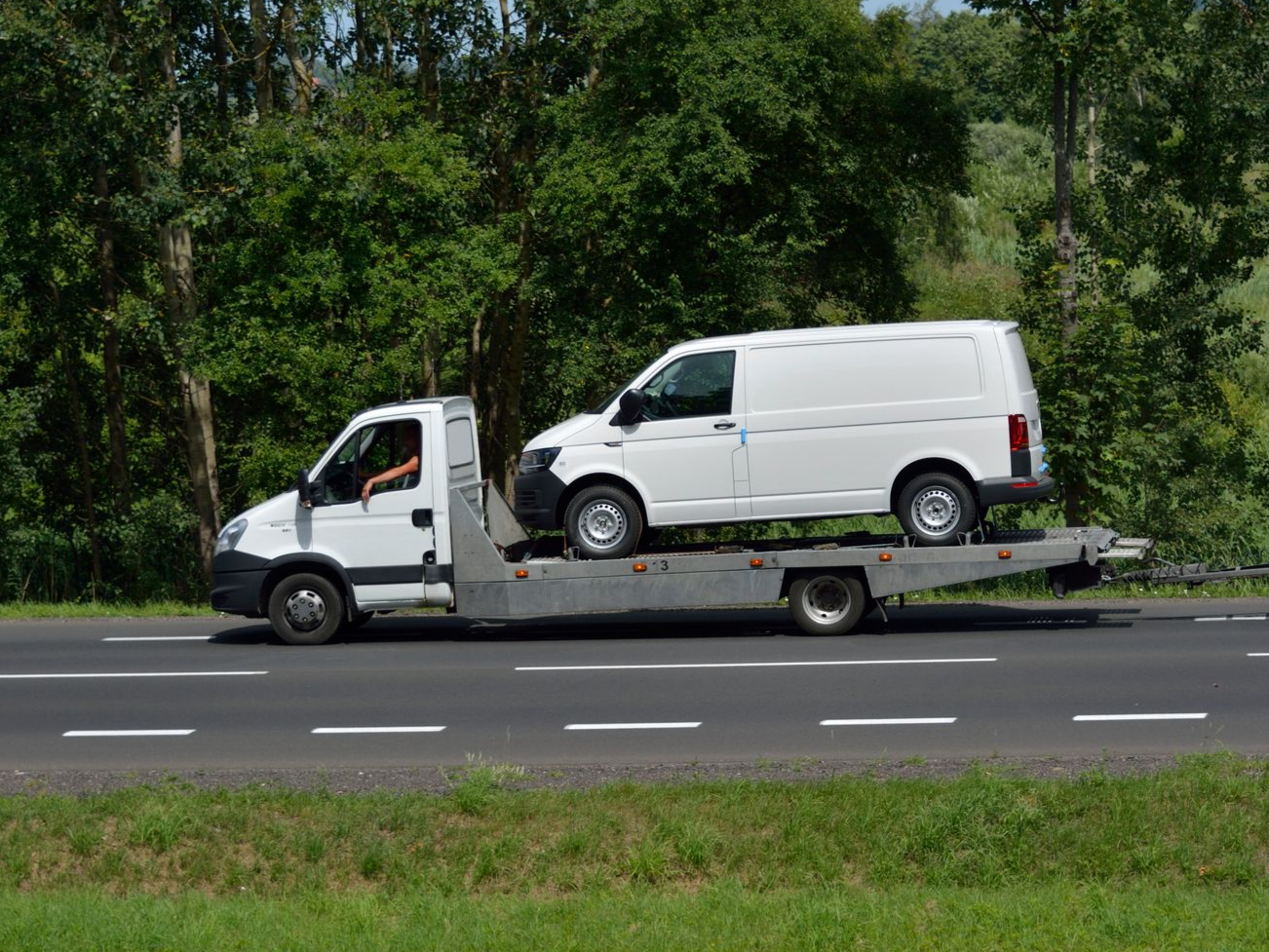 Une camionnette blanche sur une dépanneuse à plateau, en bordure de route, avec des arbres verts en arrière-plan.