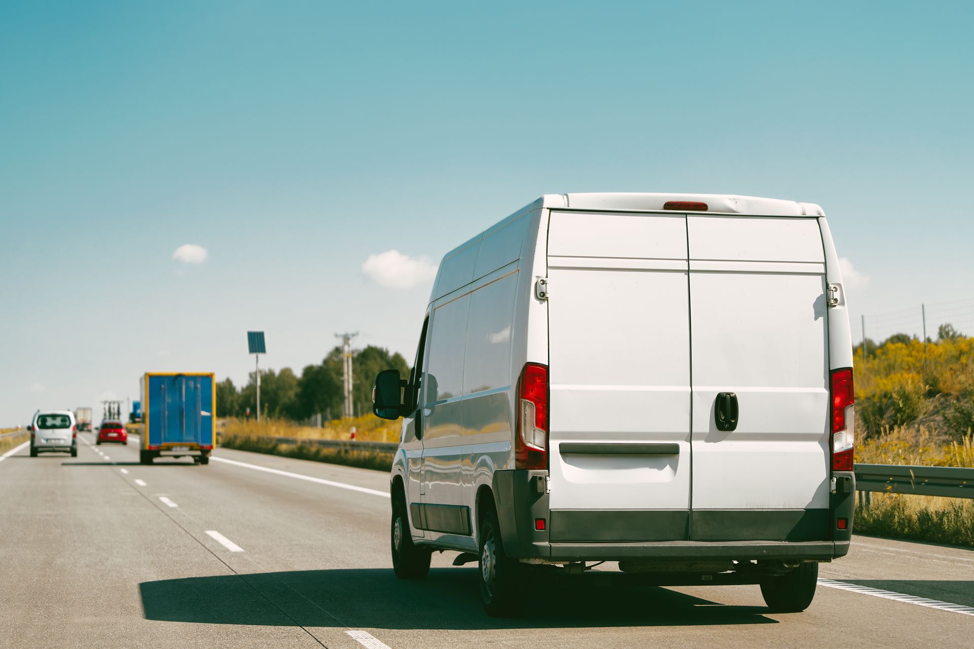 Une camionnette blanche roule sur une autoroute derrière un camion bleu sous un ciel dégagé.