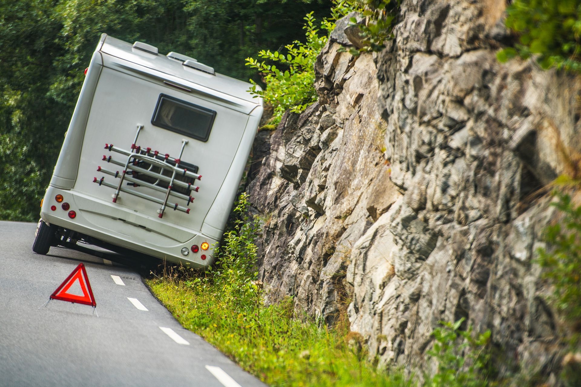 Un camping-car incliné sur une route de montagne étroite, au bord d'une falaise rocheuse, avec un triangle de signalisation rouge derrière lui.