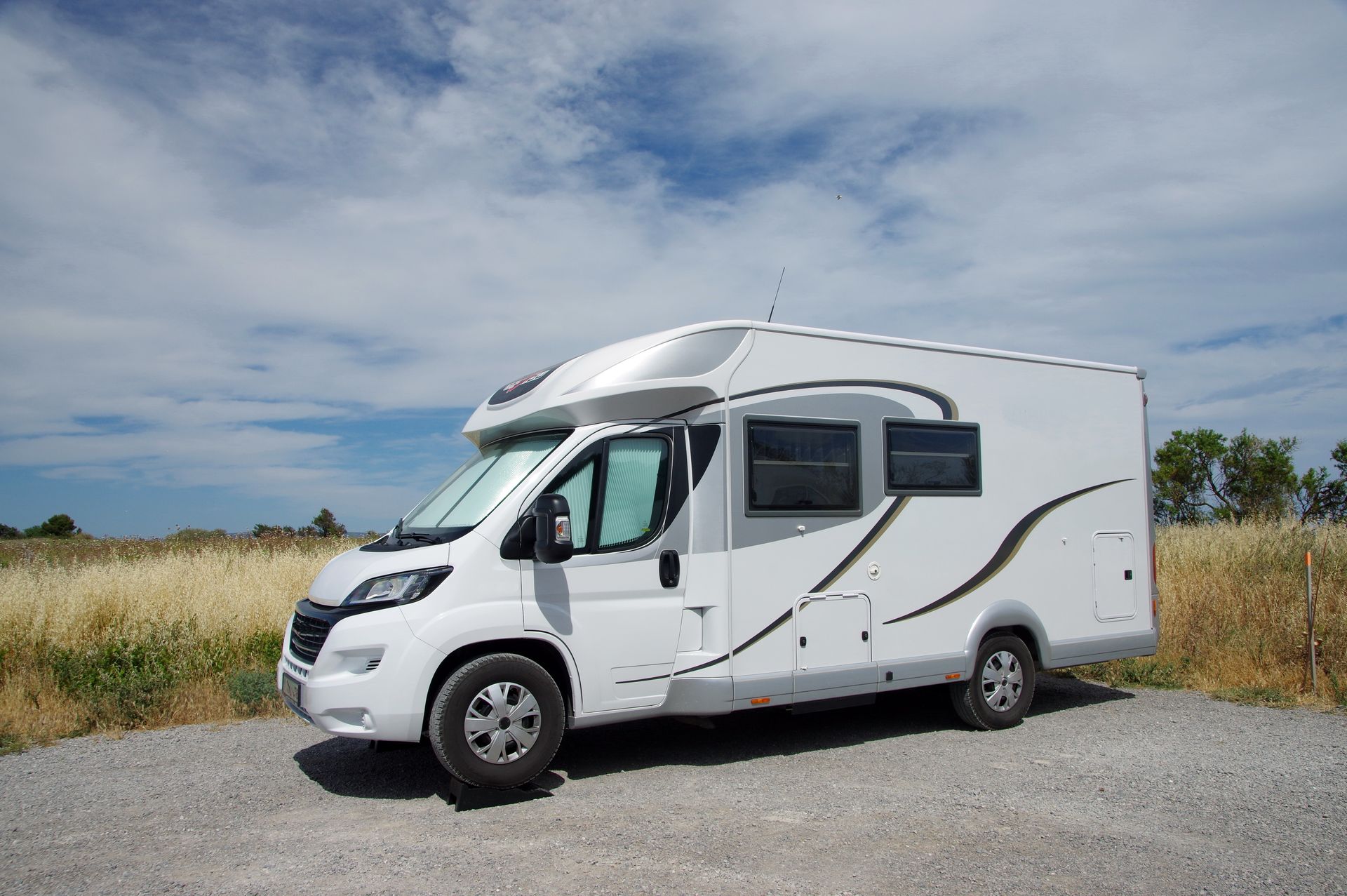 Un camping-car blanc garé sur un chemin de gravier, au bord de hautes herbes, sous un ciel nuageux.